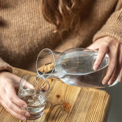 A woman in a cozy sweater pours water into a glass from a glass decanter, the concept of maintaining water balance.